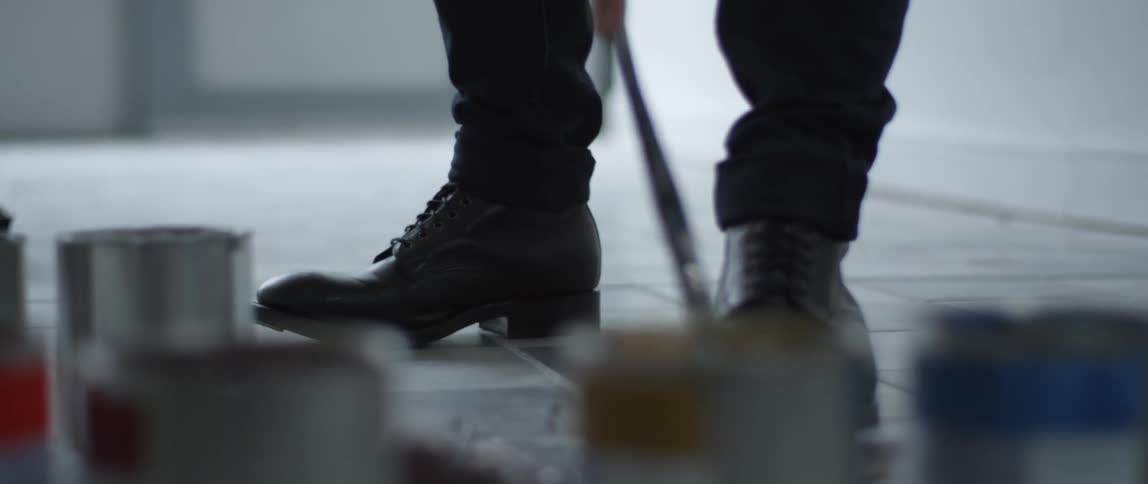 An artist wearing boots blends paint with a brush and a palette on the floor