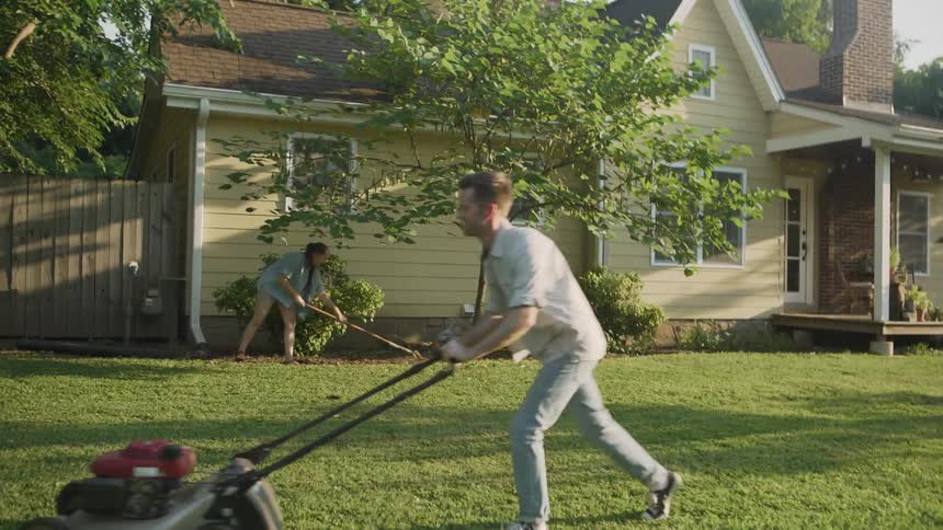 The man is pushing a lawnmower as a woman rakes
