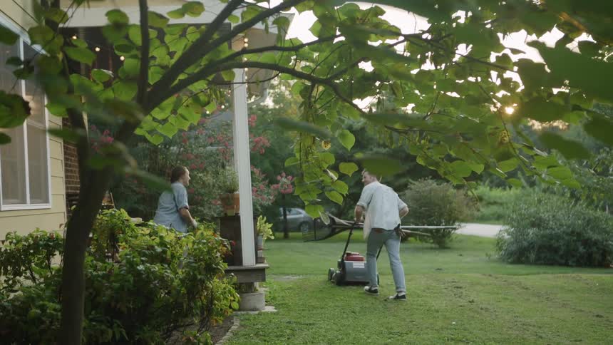 The man pushes the mower as the woman works on the plants