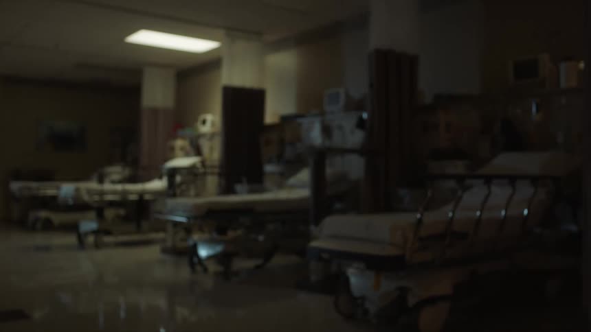 Four empty hospital beds in a dark area lit by one ceiling light