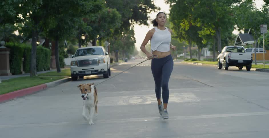 The woman is running with her dog past the cars in the neighborhood