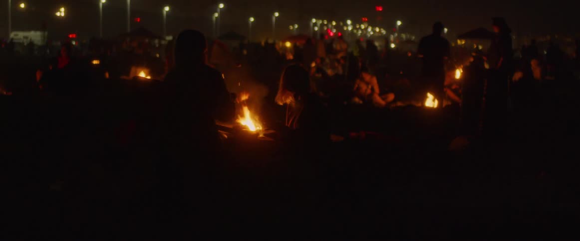 Groups of people sit around several fires on the beach at night