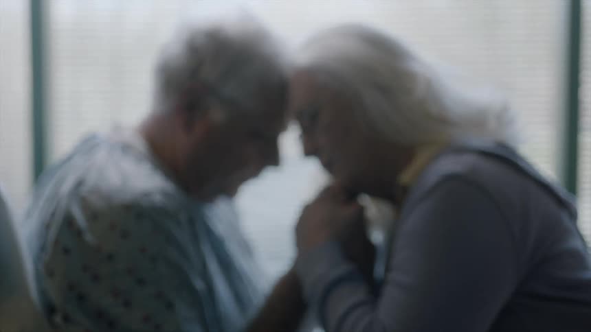 An older man wearing a hospital gown and an older woman holding hands and touching their foreheads together
