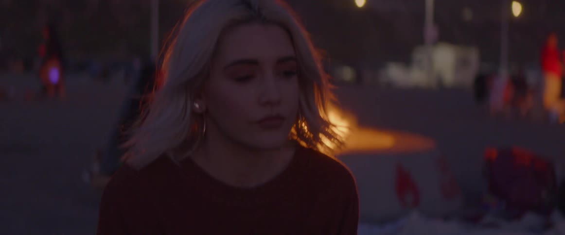 Woman with blond hair sits with her back to a bonfire under the night sky