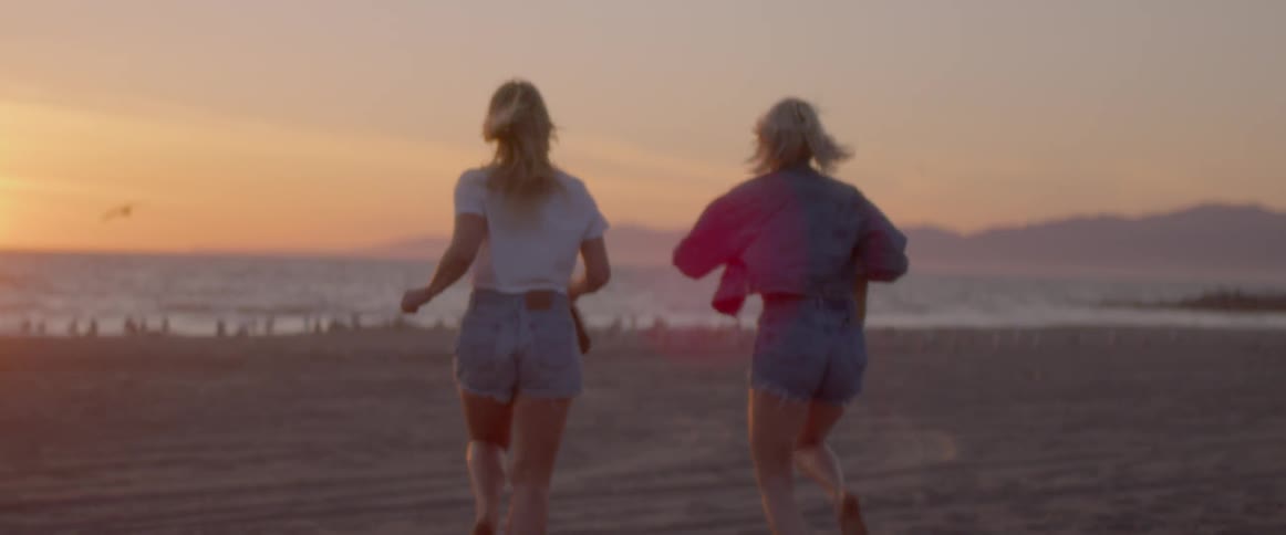 Two women holding their shoes run along a beach at sunset
