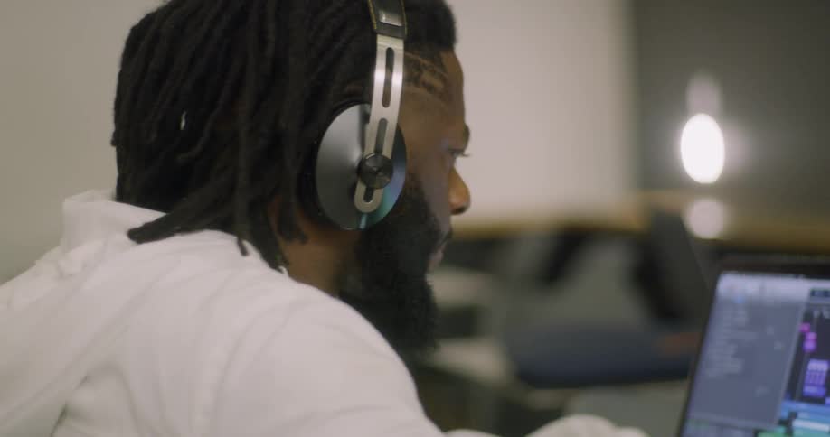 A man with dreads puts his headphones over his ears while working on a laptop