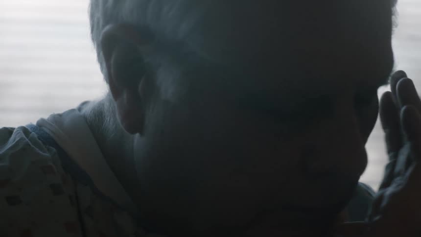 A man sits in front of a window and looks worried as he puts his head in his hand