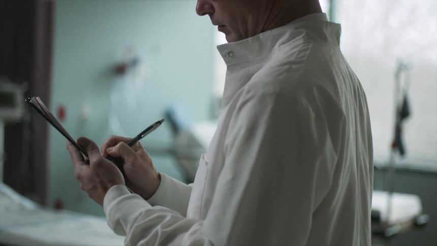 A man wearing glasses and a lab coat writing on a clipboard in a hospital room