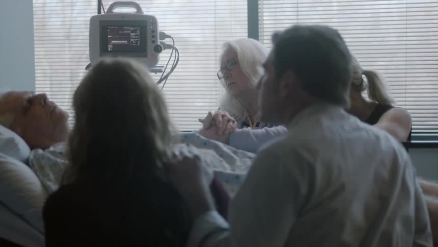 An elderly man lays in a bed in a hospital while a group of people holds hands around the bed