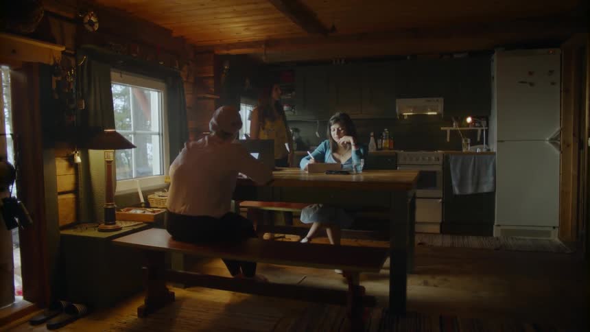 Two women sit at a table in a cabin and one woman paces behind them