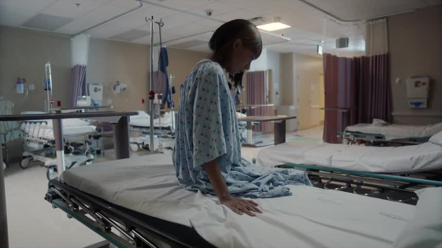 A worried black woman sits on a gurney in a hospital setting