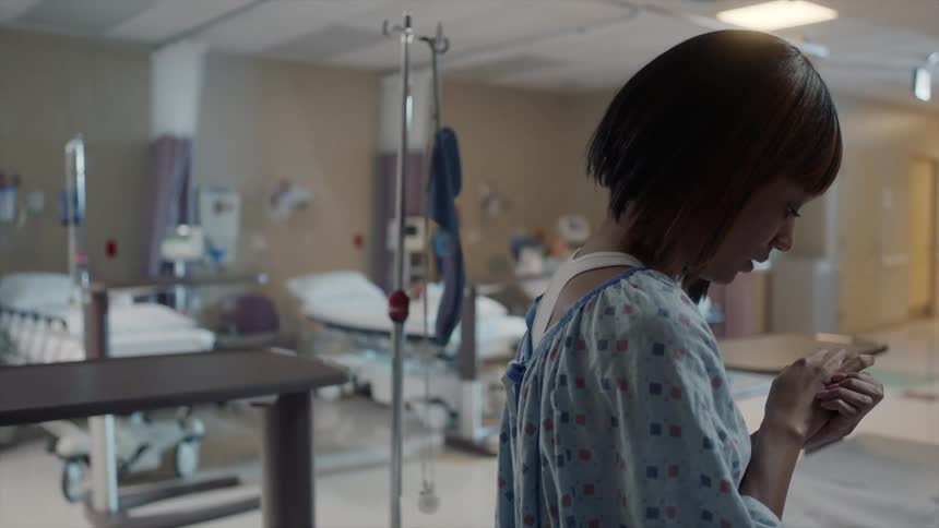 A worried woman sits on a hospital bed wringing her hands