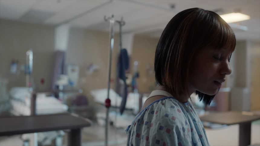 A woman with short brunette hair, wearing a hospital gown, standing in a hospital room, slowly raising her eyes to look at something before looking down again
