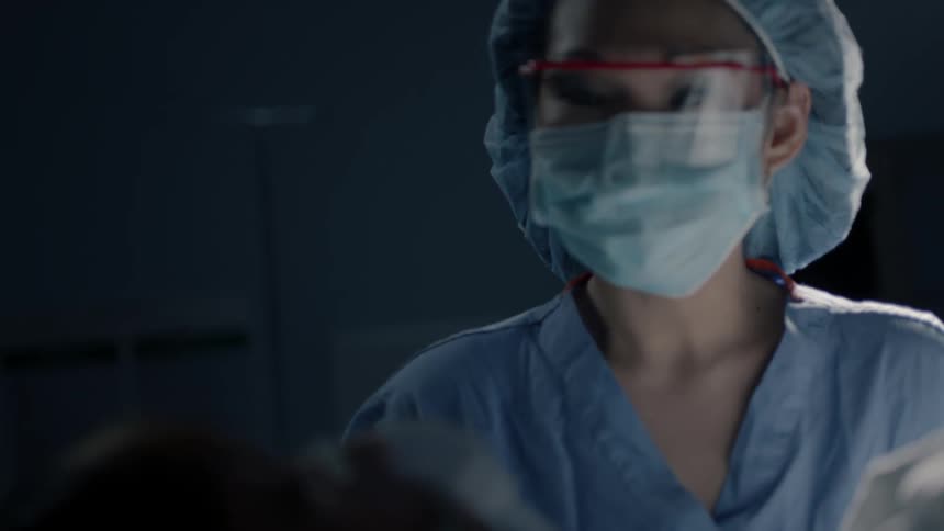 A woman in medical scrubs and a mask approaches an unconscious patient