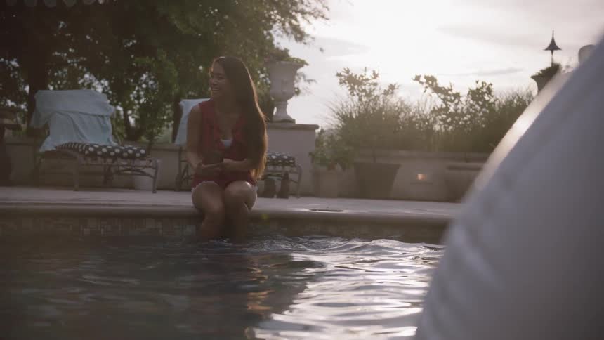 A girl sitting on the edge of a pool photographs herself with her phone while blowing a kiss