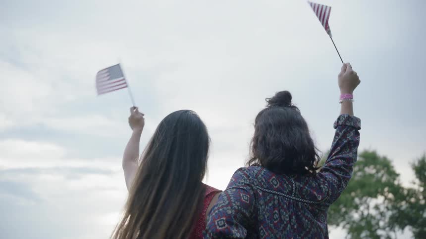 Two women are waving u.s. flags as they stand outside on a cloudy day