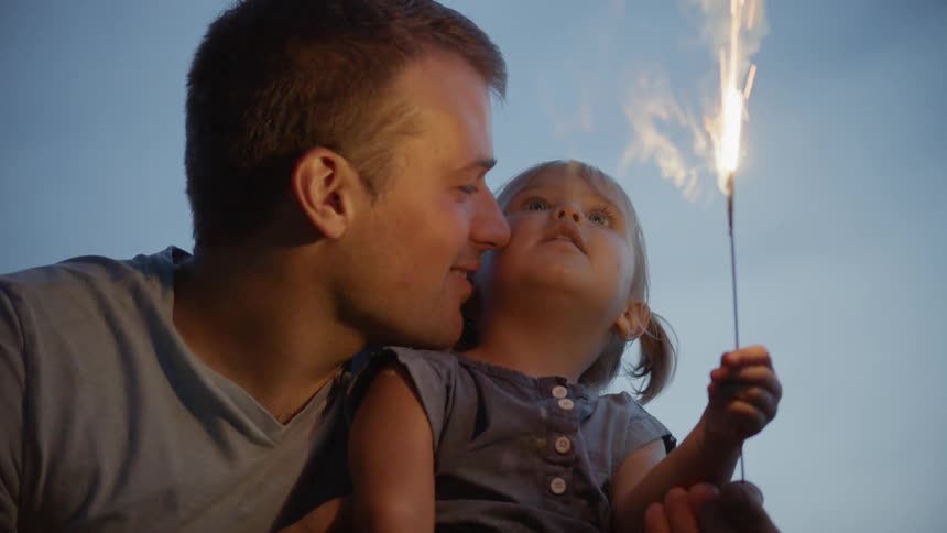 A dad smiles and snuggles his daughter who is waving a sparkler