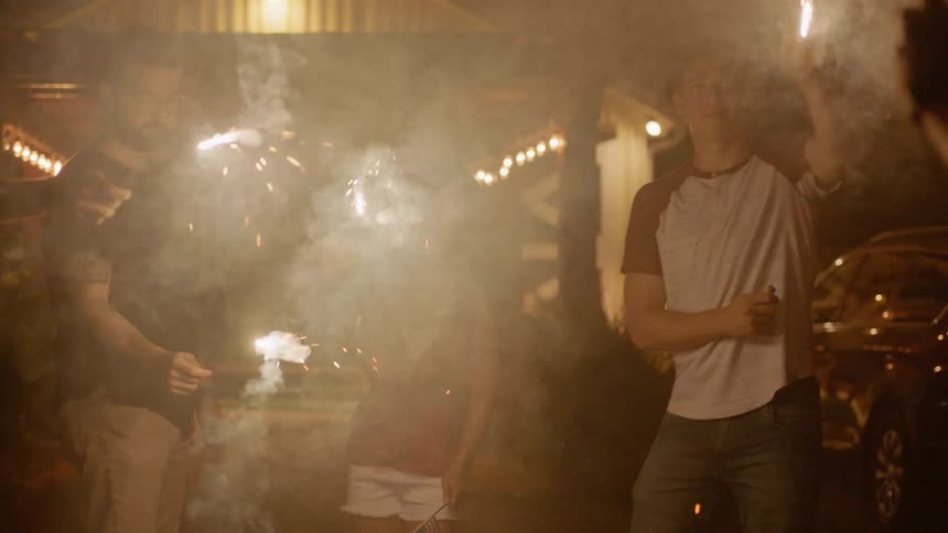 Boys and girls wave sparklers while a person records them