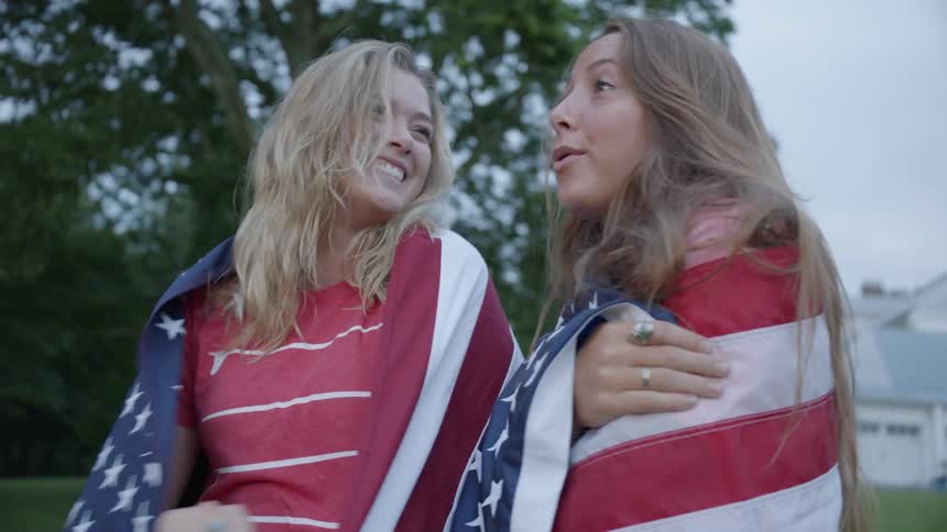 Two girls use american flags as capes as they laugh outside near trees