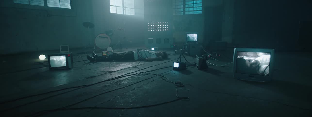 The man is lying on the floor between wires and televisions