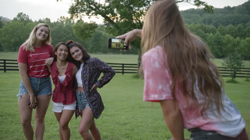 A girl uses her phone to photograph three girl friends on a grassy field
