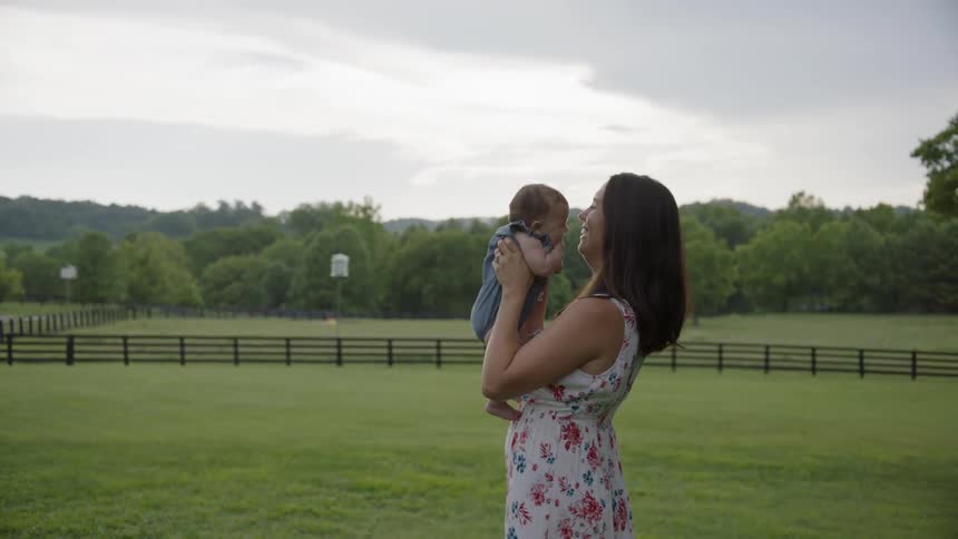 A woman holds her baby laughing and touching noses while standing in a field