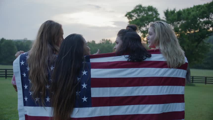 Four women are standing outside at sunset draped in an american flag