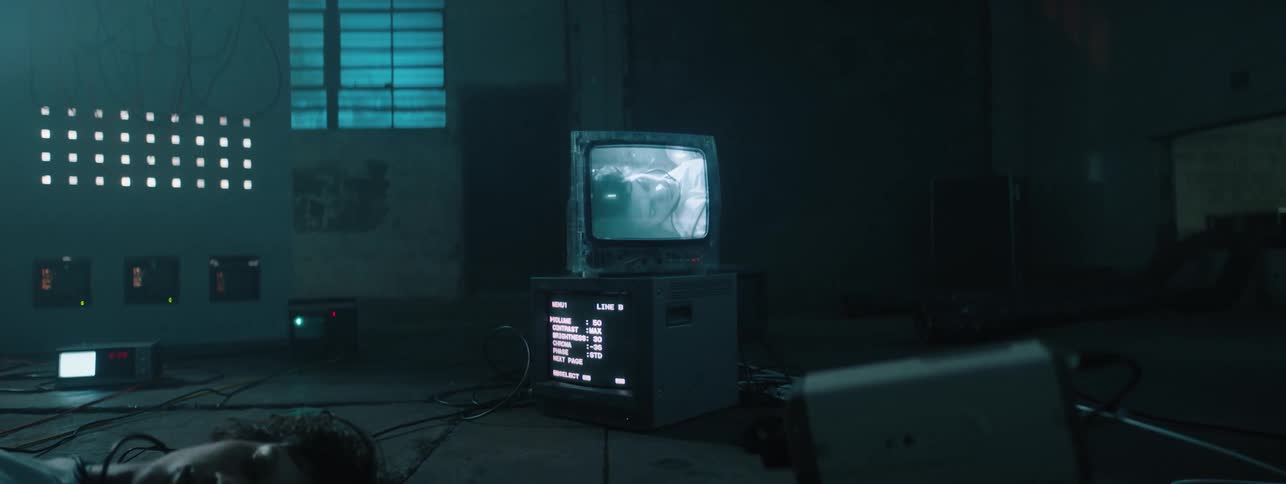 A man lays on the floor in a dark room surrounded by wires with a camera pointed at him and a monitor showing his image