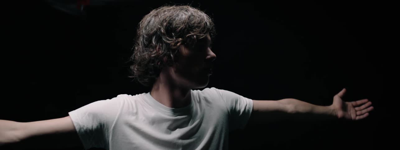 A boy in a white tee shirt stands with arms outstretched in a black room