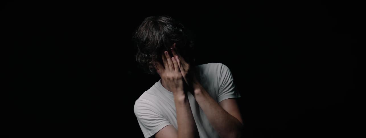 Man in a white shirt covers his face with his hands as he stands in a dark room