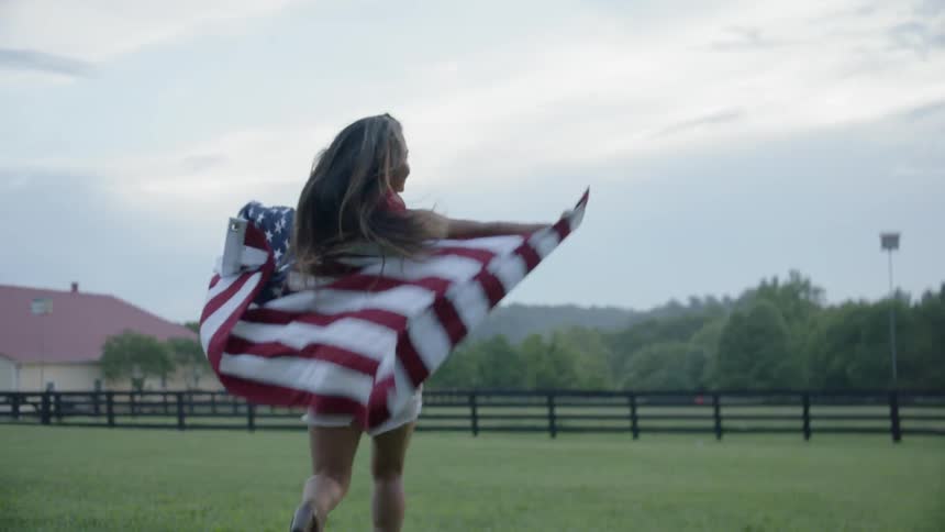 A woman runs with an american flag draped around her shoulders in a rural field