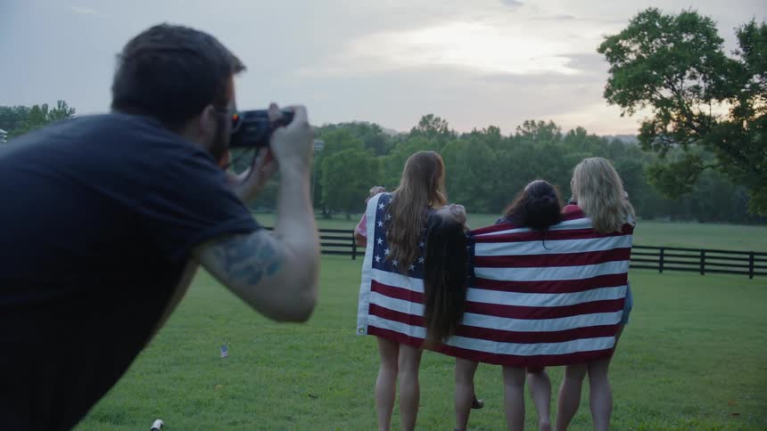 A man photographs women draped in an american flag at sunset