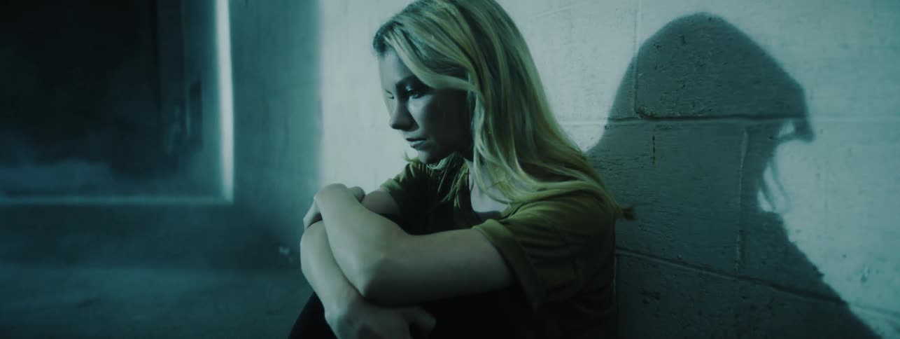 A woman sits against a cinder block wall with her arms on her knees