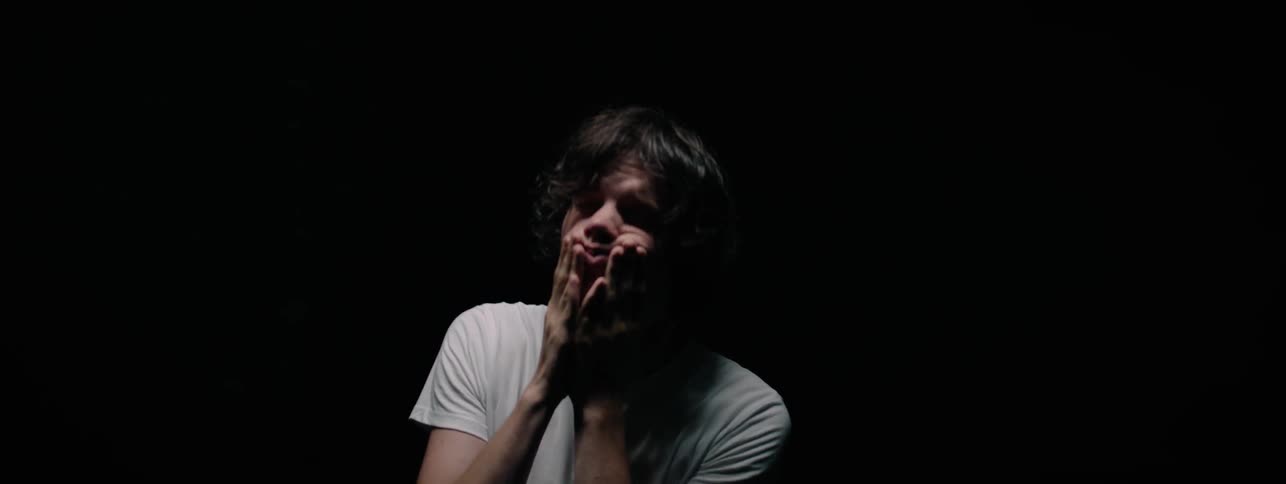 A man is rubbing and pulling at his face indoors in a dark room