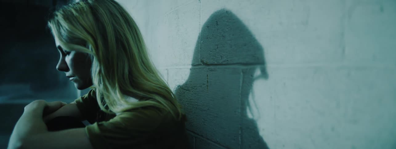 The woman sits against the wall in the dark area