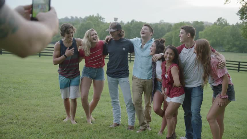 A man uses a phone to photograph a group of male and female teenage friends posing and laughing in a field