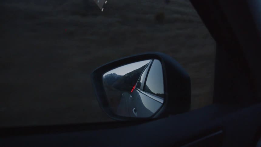 Mountains are receding in a car's mirror as it drives at dusk