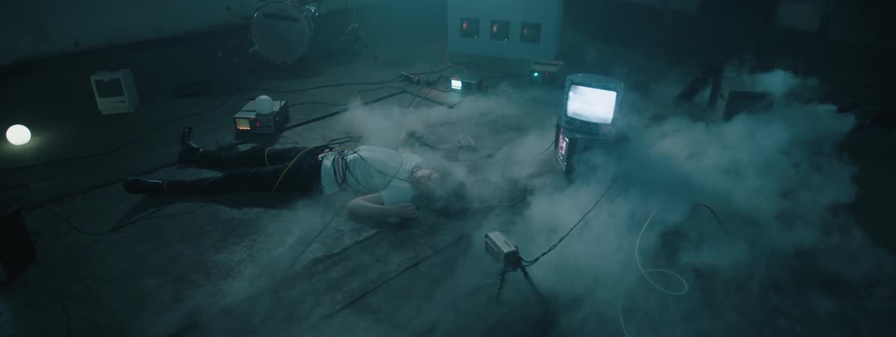 Cables and cords are wrapped around a boy lying in a room amid monitors and smoke