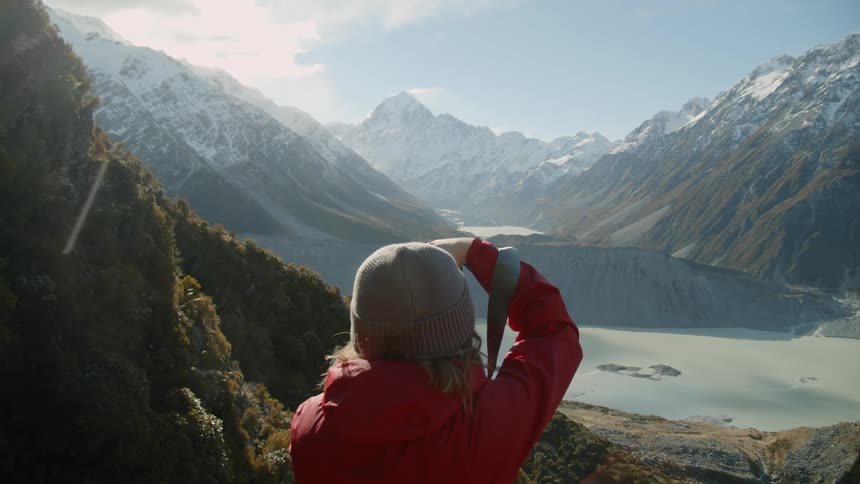 Woman taking a picture of a mountain range surrounding a half frozen lake