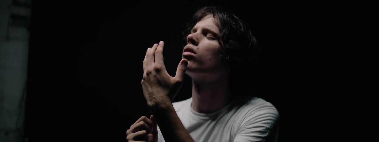 Man stands in a dark room with his eyes closed and hand raised