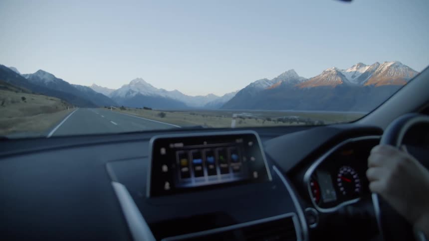 A car is being driven on a road heading towards snow-capped mountains in daylight
