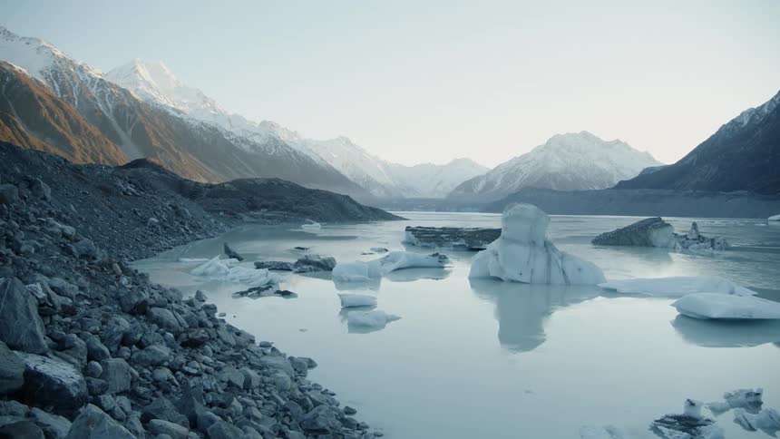 A rocky embankment leads down to a glacier filled river surrounded by mountains