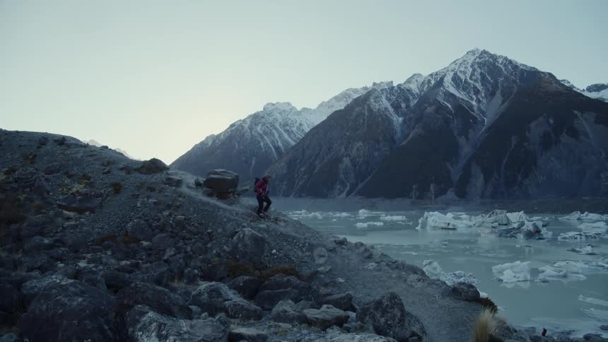 A man hikes down a path towards water with ice covered mountains in the background