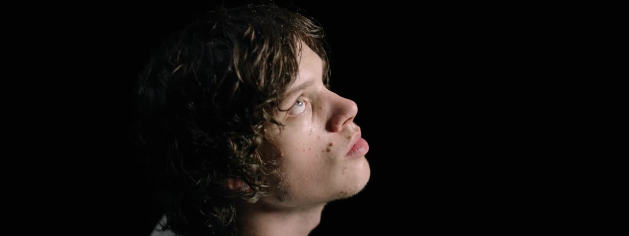 Man with black hair raises his head and looks up in a dark room