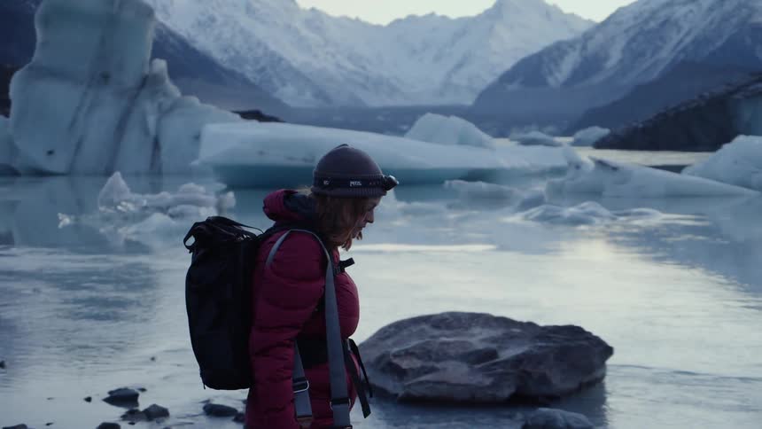 A woman wearing a backpack and carrying a camera walks on an icy shore