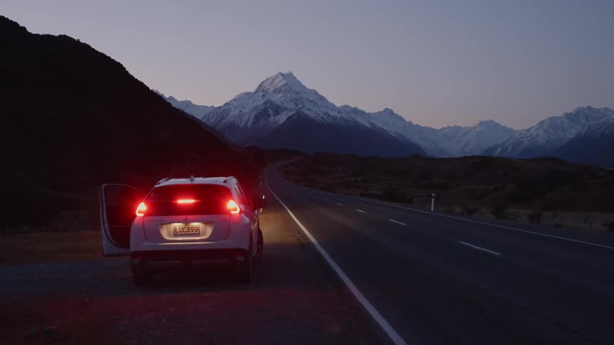 Car is parked on side of road at dusk in front of snow covered mountains