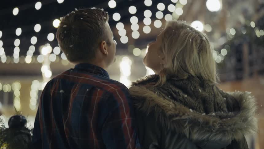 A couple stand in falling snow under lights until they kiss