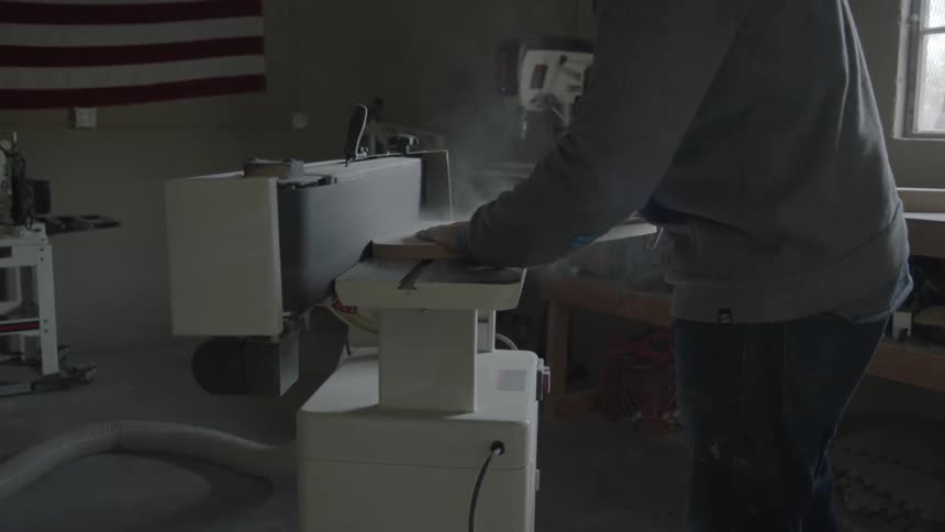 A man uses a table belt sander to shape a wooden plank in his workshop
