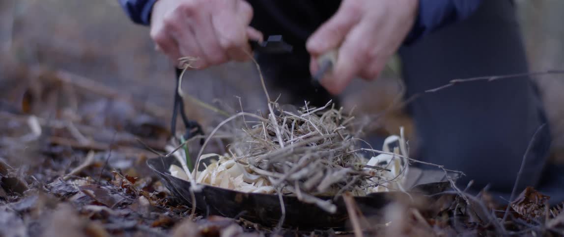 A man uses a flint to cause a spark in a pile of kindling