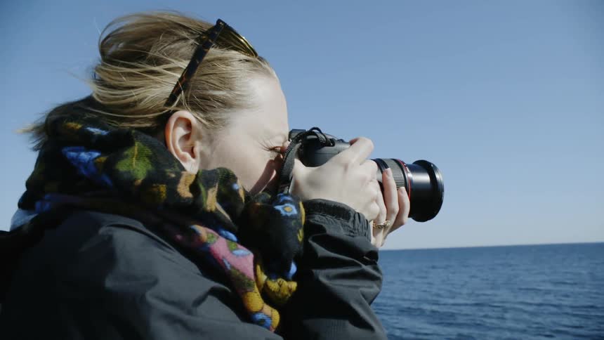 A woman looks at the ocean through her camera before bringing it away from her face and looking at the screen